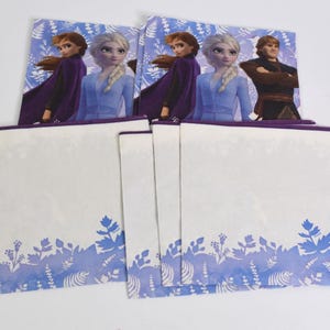 Pode incluir: Um conjunto de guardanapos para festas com o tema Frozen. Os guardanapos apresentam imagens de Elsa, Anna e Kristoff. A parte inferior dos guardanapos tem um desenho floral azul e branco. Os guardanapos s&atilde;o brancos com uma borda roxa.