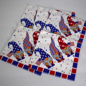 May include: A stack of white cloth napkins with a patriotic gnome design. Each napkin features a gnome wearing a blue star-covered hat or a red and white striped hat, holding an American flag. The napkins have a red and blue border.