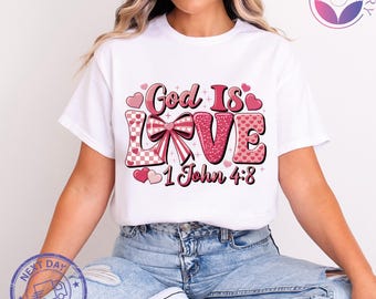 May include: White t-shirt with a pink and red graphic that reads "God Is Love 1 John 4:8". The design includes hearts and a bow. The shirt is a crew neck style.