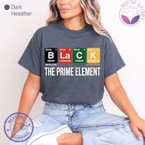 May include: Dark heather gray t-shirt with the word "BLACK" spelled out using periodic table elements. The shirt also features the text "THE PRIME ELEMENT" in white. The shirt is a casual style.