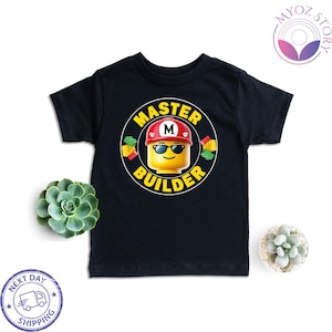May include: Black short-sleeve t-shirt featuring a graphic of a Lego figure wearing sunglasses and a red hard hat with the letter "M". The text "MASTER BUILDER" is in a yellow circle around the figure. The shirt is for children.
