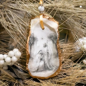 Può includere: Un ornamento natalizio realizzato con un guscio di ostrica naturale, con una scena della Natività dipinta a mano in bianco e nero. Il guscio ha un bordo dorato ed è sospeso da un nastro dorato con una perla decorativa. L'ornamento è su un albero di Natale.