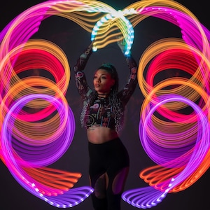 May include: A person in black leggings and a patterned crop top is surrounded by vibrant, glowing light trails in shades of purple, orange, and pink. They hold a glowing, curved light above their head. The image is set against a dark background.