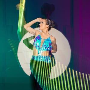 May include: A young woman wearing a blue and silver sequined top and black pants poses in a spotlight. The background is a dark blue and purple gradient with green and yellow light streaks.
