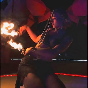 Puede incluir: Una persona actuando con fuego. La persona lleva un atuendo negro y sostiene objetos en llamas. El fondo es oscuro con acentos rojos y naranjas, creando una escena dramática y enérgica.