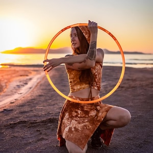 May include: A person wearing a brown tie-dye top and skirt is kneeling on a sandy beach at sunset. They are holding a large orange hoop in front of them.