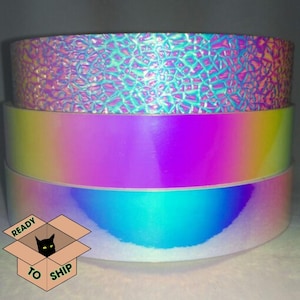 May include: Three rolls of iridescent decorative tape in a stack. The top roll has a textured, cracked-glass appearance, while the middle roll transitions from yellow to purple. The bottom roll reflects blue and green hues. A box with a cat and the words "READY TO SHIP" is in the lower left.