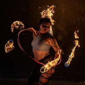 May include: A woman performs a fire hoop dance with flaming torches. She is wearing a striped crop top and black pants. The background is dark with a wall behind her.