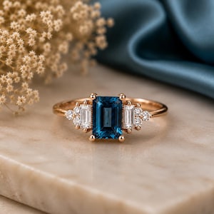Op de afbeelding: Een ring van roségoud met een centrale smaragdgeslepen donkerblauwe edelsteen, geflankeerd door twee heldere stenen in baguette-slijpvorm en kleinere ronde heldere stenen aan weerszijden. De ringband is gemaakt van gepolijst roségoud.