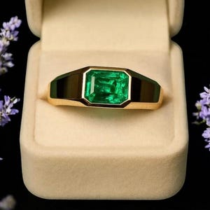Op de afbeelding: Een gouden ring met een grote, rechthoekige smaragd. De smaragd is heldergroen en gezet in een gouden band met zwarte accenten. De ring wordt gepresenteerd in een beige ringdoos, met paarse bloemen op de achtergrond.
