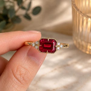 May include: A gold ring featuring a rectangular emerald-cut red gemstone at the centre, framed by a beaded edge. The setting includes three small round white accent stones on each side of the central gem, mounted on a thin, polished gold band.