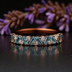 May include: A rose gold ring featuring alternating rectangular blue gemstones and small clear stones. The ring is set against a blurred background of purple flowers.