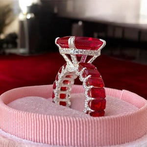 May include: A close-up of a ruby ring set in a pink velvet ring box. The ring features a large, rectangular ruby center stone and smaller rubies set along the band. The ring is surrounded by small, clear accent stones.