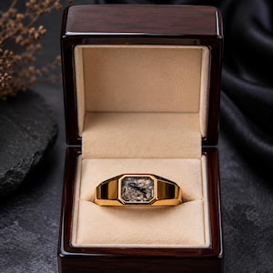 May include: A gold signet ring featuring a rectangular rutilated quartz gemstone with visible black needle-like inclusions, presented inside a polished dark wood jewelry box with a beige velvet interior lining and a hinged lid.
