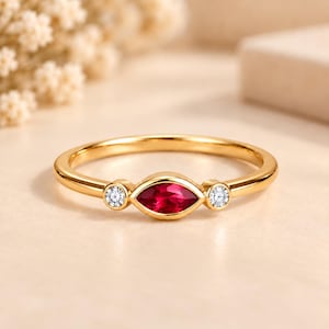 Op de afbeelding: Een gouden ring met een centrale robijnrode edelsteen in marquise-slijpvorm, geflankeerd door twee kleine, ronde, helderwitte steentjes. De ringband is glad en gepolijst, met een strak ontwerp en een zetting voor elk van de drie stenen.