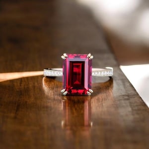 May include: A gold ring with a large, rectangular, deep red gemstone as the focal point. The band is adorned with small, clear stones. The ring is set against a dark, wooden surface, creating a contrast that highlights the jewelry.
