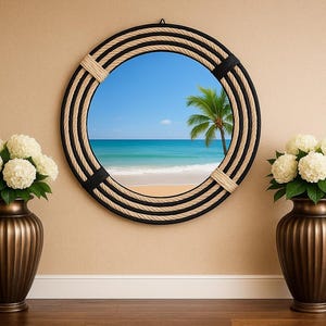 May include: A round mirror with a rope and black trim, showcasing a beach scene with a palm tree. Flanking the mirror are two bronze-colored vases filled with white flowers, set against a beige wall and wooden floor.