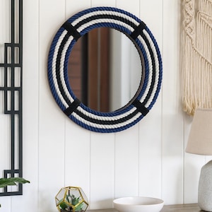 May include: Round mirror with a nautical design. The frame is wrapped with blue, white, and black rope. Black bands secure the rope. The mirror hangs on a white wall. A wooden cabinet with a terrarium and bowl is below.