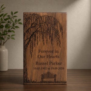 Könnte beinhalten: Rechteckige Urne aus Holz mit Gravur einer Trauerweide. Der Text "Forever in Our Hearts" und der Name "Russel Parker" sind unter dem Baum zusammen mit Daten eingraviert. Am unteren Rand ist eine Bank eingraviert.
