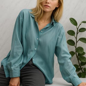 Puede incluir: Una camisa abotonada de color verde azulado con mangas largas. La camisa está desabrochada en la parte superior y combinada con pantalones grises oscuros. La camisa tiene un corte relajado y está hecha de una tela suave y fluida.