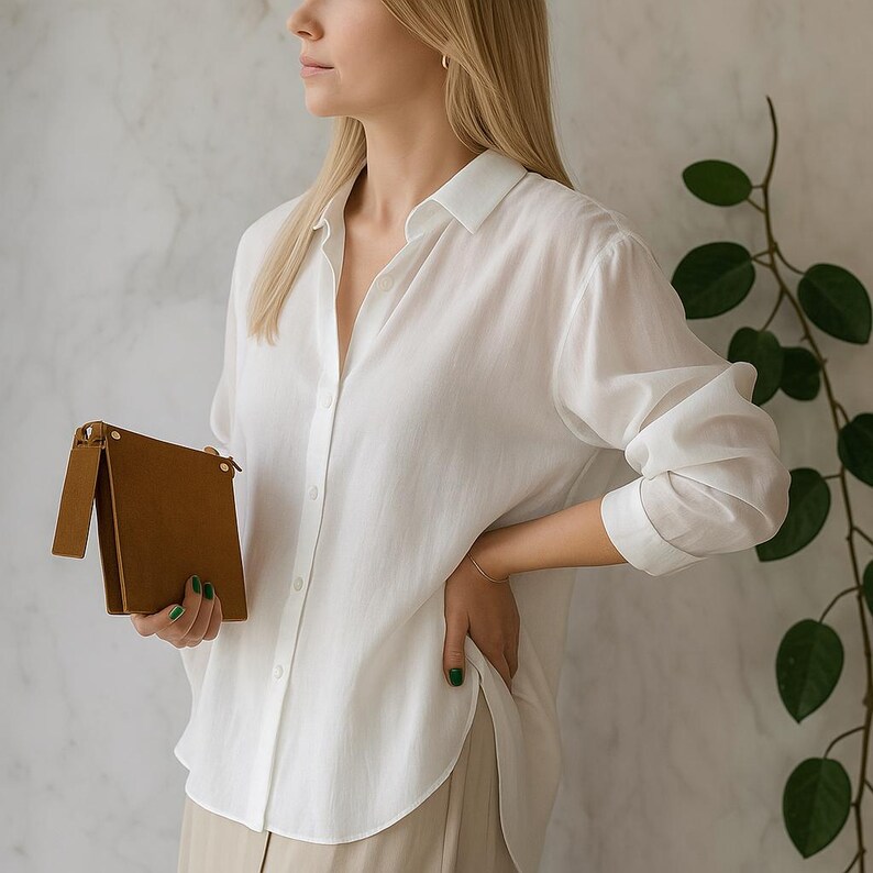 May include: A long-sleeved white button-down shirt with a classic collar and a curved hemline, paired with a small, rectangular tan leather clutch bag featuring a fold-over flap and gold-toned metal hardware accents.
