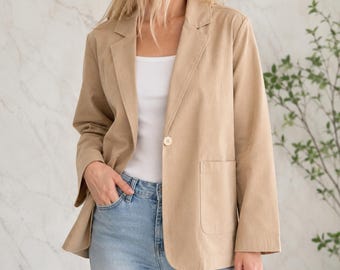 May include: A tan blazer featuring a single-button closure, notched lapels, and two large patch pockets on the front. The blazer is worn over a white tank top and light blue denim jeans, creating a neutral and layered appearance.