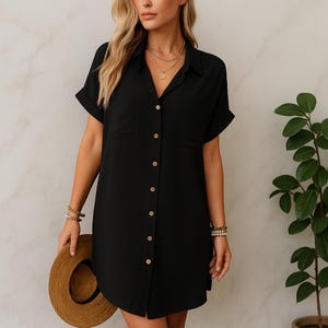 May include: Black shirt dress with short sleeves and a collared neckline. The dress has a single chest pocket and wooden buttons. A straw hat is held in the hand. The dress is knee-length.
