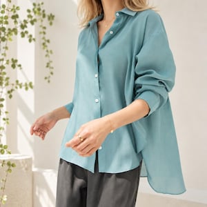 May include: A long-sleeved button-down shirt in a muted teal color, featuring a classic collar, a front button placket, and a slightly longer back hem. The shirt is paired with dark gray trousers, creating a simple and clean ensemble.