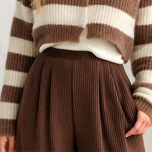May include: A brown and cream striped cardigan with a white undershirt and brown corduroy pants. The cardigan has a button closure. The pants are high-waisted and pleated. The outfit is a cozy, fall-themed ensemble.