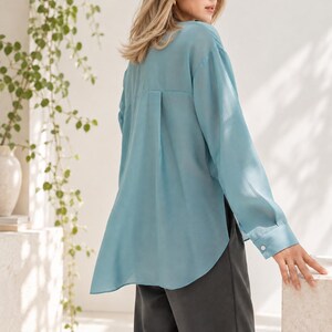 May include: A long-sleeved, light blue button-down shirt with a semi-sheer finish, a back yoke, a center box pleat, and a curved high-low hemline, paired with dark gray trousers. The shirt features cuffed sleeves with a single white button.
