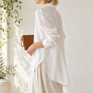 May include: A long-sleeved white button-down shirt with a collar and a curved hemline, paired with a light-colored skirt. A tan leather clutch bag is held in one hand, featuring a wrist strap and a simple rectangular design.