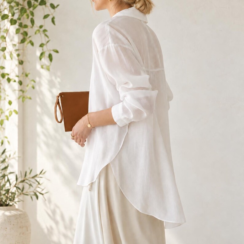 May include: A long-sleeved white button-down shirt with a collar and a curved hemline, paired with a light-colored skirt. A tan leather clutch bag is held in one hand, featuring a wrist strap and a simple rectangular design.