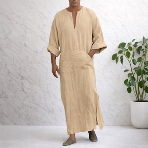 May include: A beige, long-sleeved robe with a V-neck and a front pocket. The robe is floor-length and has side slits. The sleeves are rolled up at the elbows. The fabric appears to be linen or a similar lightweight material.