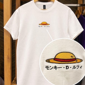 Op de afbeelding: Wit t-shirt met een geel en rood strohoedontwerp en Japanse tekst. Het hoedontwerp is op de voorkant geborduurd. Het t-shirt hangt aan een houten hanger.
