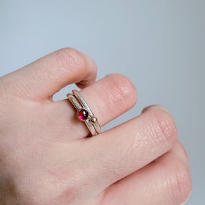 May include: A stack of two silver rings worn on a finger. One ring features a small, round, deep red gemstone set in a bezel, while the other ring is a simple silver band accented with a small, smooth, polished gold-toned metal sphere.