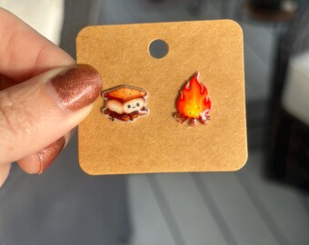 May include: A pair of novelty earrings on a brown card. One earring is a cartoon s'more with a smiling face. The other earring is a cartoon campfire with red, orange, and yellow flames. The earrings are held by a person.