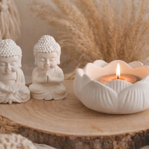 Könnte beinhalten: Zwei weiße Buddha-Statuen und ein weißer Lotusblüten-Kerzenhalter mit einer brennenden Kerze sitzen auf einer Holzscheibe. Die Statuen befinden sich in einer sitzenden Meditationshaltung. Der Kerzenhalter hat eine einzelne brennende Kerze.