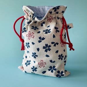 May include: A drawstring bag with a floral pattern. The bag is cream-colored with a repeating pattern of blue and red flowers. It has a red drawstring closure and a light blue interior.