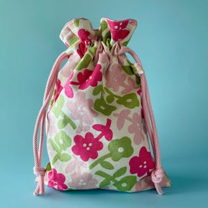May include: A drawstring bag with a floral pattern in pink, green, and white. The bag is tied with a pink cord. The flowers are in a repeating pattern. The background is a solid light blue.