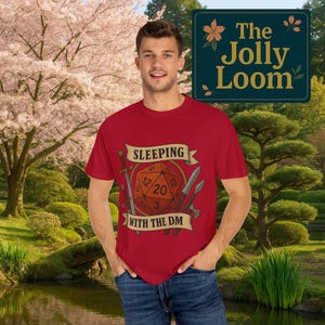 May include: A red t-shirt with the text "SLEEPING WITH THE DM" and a graphic of a 20-sided die, swords, and knives. The Jolly Loom logo is in the upper right corner. The person is wearing blue jeans.