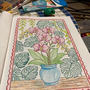 May include: A detailed coloring page featuring a blue vase with pink orchids and green leaves. The artwork is surrounded by large, stylized leaves and a decorative pink border. The page is open on a table with art supplies.