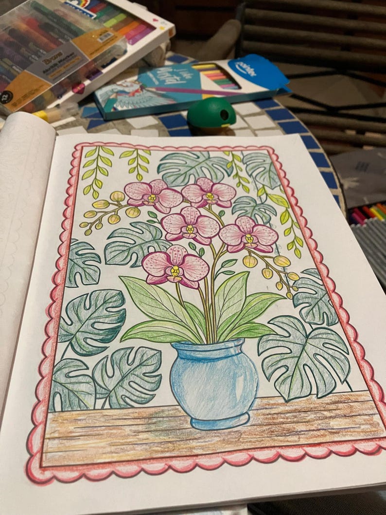 May include: A detailed coloring page featuring a blue vase with pink orchids and green leaves. The artwork is surrounded by large, stylized leaves and a decorative pink border. The page is open on a table with art supplies.