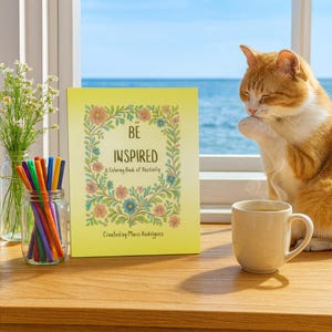 May include: A yellow coloring book titled "BE INSPIRED" with floral designs and the text "& Coloring Book of Positivity." A mug, a jar of colored pencils, and a vase of flowers sit on a wooden surface. An orange and white cat is in the background.