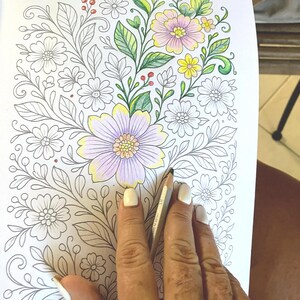 May include: A partially colored floral coloring book page with a hand holding a colored pencil. The flowers are colored in shades of pink, yellow, and purple, with green leaves and red berries. The rest of the flowers are uncolored.