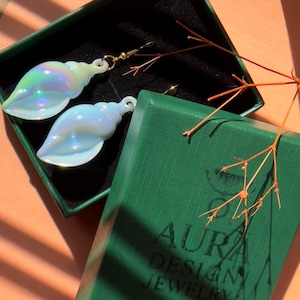 May include: Iridescent seashell earrings with gold-colored hooks, presented in a green jewelry box. The earrings have a pearlescent sheen, reflecting various colors. The box has the text "AURA DESIGN JEWELRY" printed on it.