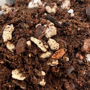 May include: Close-up of a pile of potting soil mix, featuring dark brown soil, pieces of bark, and white perlite. The mix appears to be a blend of organic materials, suitable for planting.