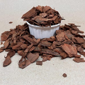 Puede incluir: Un cuenco de cerámica blanco rebosante de mantillo de corteza marrón rojizo, rodeado de un montón disperso del mismo material. Las piezas de corteza varían en tamaño y forma, creando una apariencia natural y texturizada. Ideal para jardinería y paisajismo.