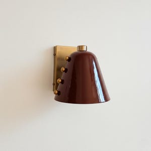 May include: A wall-mounted sconce with a dark red, bell-shaped shade and a brass-colored mounting plate. The sconce features decorative brass-colored rivets and a small, rounded top. The design is modern and minimalist.