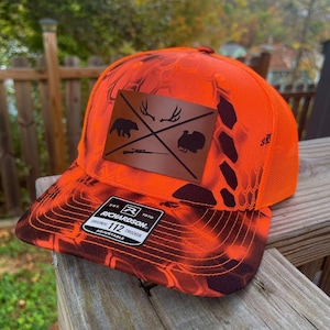 May include: An orange and black trucker hat with a brown leather patch featuring a bear, antlers, crossed rifles, and a turkey. The hat has a mesh back and the text "RICHARDSON" on the brim.