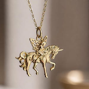 May include: A gold-toned metal pendant necklace featuring a detailed figure with wings riding a unicorn. The pendant is suspended from a delicate gold-toned chain. The design shows the winged figure seated on the back of the unicorn, which is depicted in a standing position with its horn clearly visible.